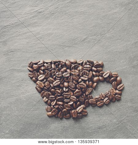 Cup from coffee beans. Heap of roasted coffee bean on grey stone surface texture. Coffee shop or cafe background. Natural stone and cup from beans. Soft color toning, top view