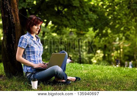 Beautiful young freelancer woman using laptop sitting under the tree in the park.Happy smiling girl working online.Studying and learning using notebook computer.Freelance workbusiness people concept.