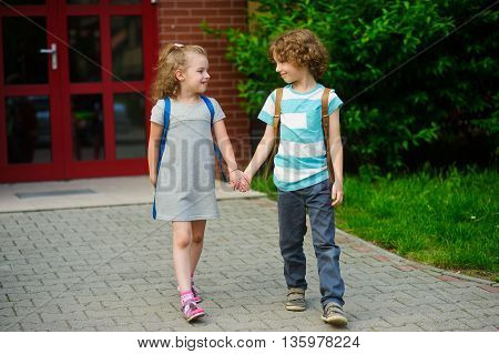 Little pupils on a schoolyard. The boy and the girl go having joined hands. Behind shoulders at school students satchels. Children look at each other and smile. Back to school.