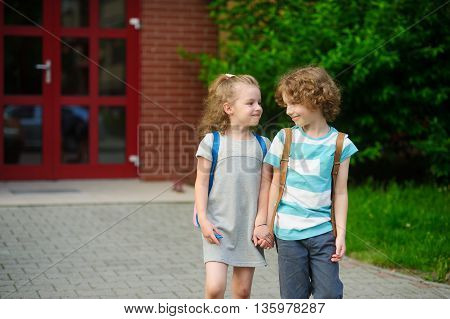 Little schoolmates on a schoolyard. The boy and the girl go having joined hands. Behind shoulders at school students satchels. Children look at each other and smile.