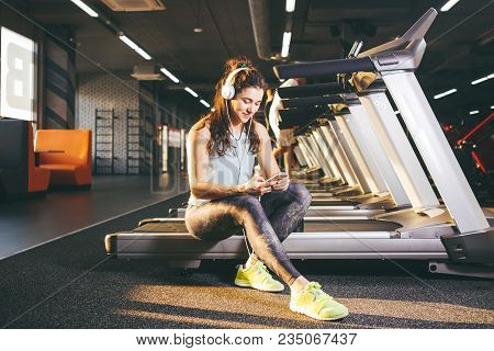 Beautiful Young Caucasian Girl Sportswoman Sitting, Resting After Training On Treadmill Against The 
