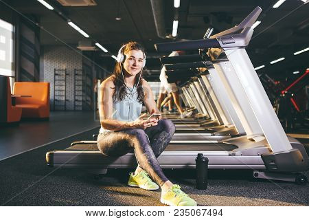 Beautiful Young Caucasian Girl Sportswoman Sitting, Resting After Training On Treadmill Against The 