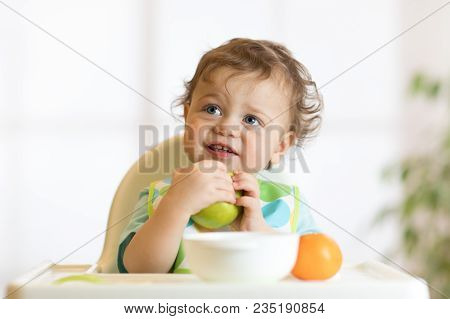 Smiling Little Kid Child Baby Boy Toddler Sitting In Highchair And Eating Big Green Apple Fruit Port
