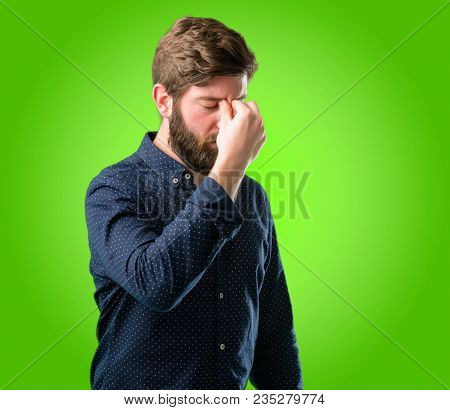 Young hipster man with big beard with sleepy expression, being overworked and tired, rubbes nose because of weariness over green background