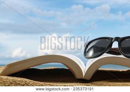 Close Up Book And Glasses On Sand Of Beach On Blur Beautiful Sky Background, Summer Concept