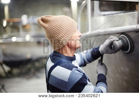 Mature worker of shipbuilding factory using worktools in process of constructing new boat