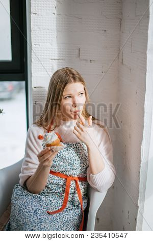 Young Woman Enjoys Her Cupcake Sitting Against White Wall , Licking The Cream From Her Finger Lookin