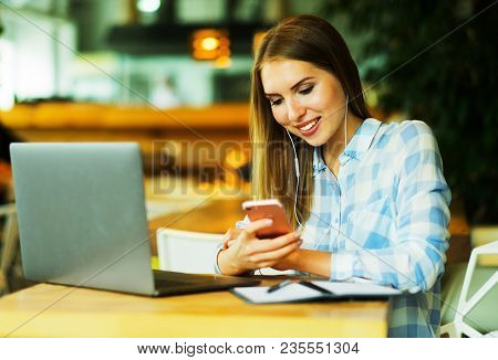 Young beautiful blond girl sitting in a coffee shop and working on computer. Image of happy woman using laptop and smartphone in cafe.