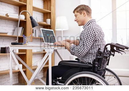 Daily Lifestyle. Positive Delighted Man Using His Computer While Sitting On Wheelchair