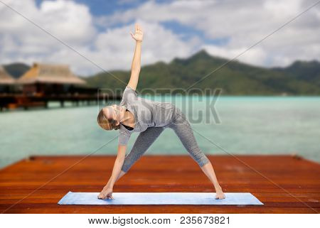 fitness, sport, people and healthy lifestyle concept - woman making yoga triangle pose on wooden pier over island beach and bungalow background