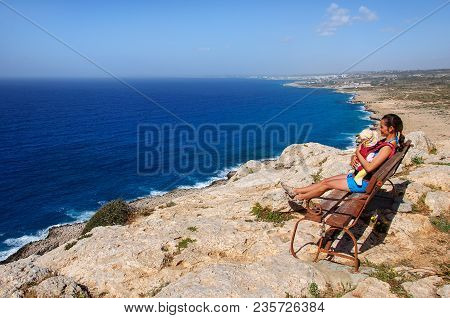 Young Woman With A Child Sitting On A Bench On A Rocky Shore Of The Mediterranean Sea, Cyprus