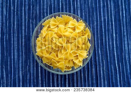 Farfalle Raw Pasta In A Glass Bowl On A Striped White Blue Fabric Background In The Center. Close-up