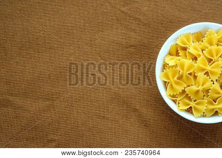 Farfalle Raw Pasta In A White Bowl On A Brown Rustic Background Texture With A Side. Close-up With T