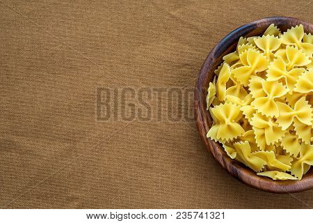 Farfalle Raw Pasta In A Wooden Bowl On A Brown Rustic Background Texture With A Side. Close-up With 
