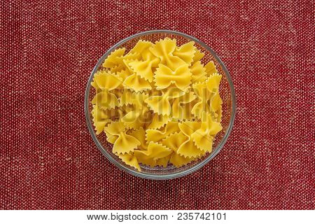 Farfalle Raw Pasta In A Glass Bowl On A Red Brown Rustic Texture Background, In The Center Close-up 