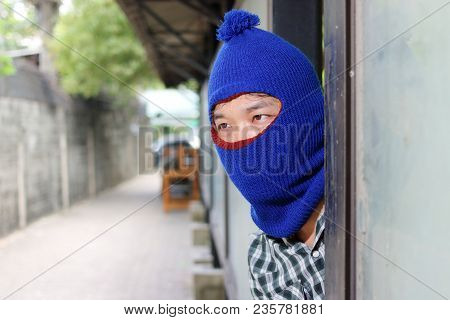Masked Burglar Wearing A Balaclava Sneaking Behind A Pole Before The Burglary. Crime Concept