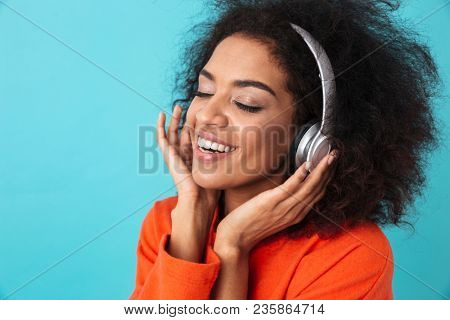 African american glad woman in orange shirt listening to music via headphones with smile isolated over blue background