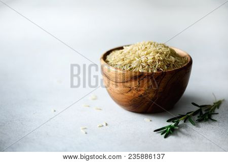 Raw Organic Brown Rice In Wooden Bowl And Rosemary On Light Concrete Background. Food Ingredients. C
