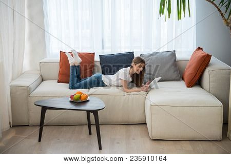Side View Of Young Female Lying On Couch And Using Digital Tablet In Living Room With Modern Interio