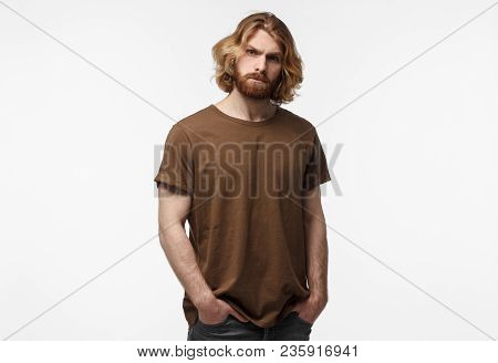 Young Brutal Bearded Man With Long Hair Standing With Hands In Pockets, In Brown Tshirt, Isolated On