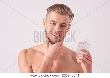 Attractive young man applying aftershave lotion on face and smiling while standing isolated on white background. Cheerful bearded hipster holding after shave cream bottle in hand and touching chin