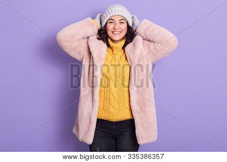 Horizontal Indoor Picture Of Joyful Adorable Young Woman Putting On Her Light Hat, Looking Directly 