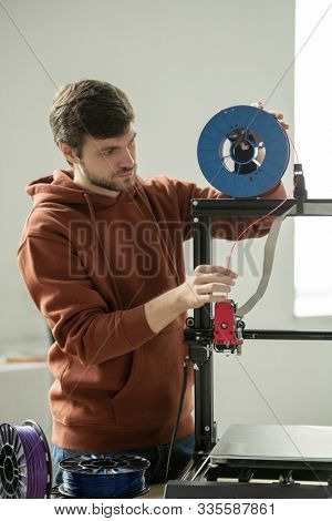 Young man in casualwear putting red filament into printhead of 3d printer before printing new items
