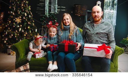 Christmas Concept - Happu Family Sitting On The Couch Holding Gifts And Looking In The Camera