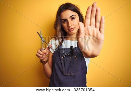 Young beautiful hairdresser woman holding scissors over yellow isolated background with open hand doing stop sign with serious and confident expression, defense gesture