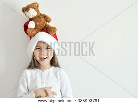 Smiling Little Girl In A Christmas Santa Hat With A Toy Teddy Bear On The Head. Child Isolated On A 