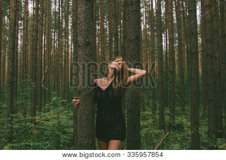 Portrait Of Young Beautiful Girl In Black Dress With Threw Back Head In Forest That Holds Tree Trunk