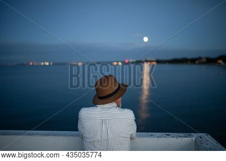 Rear View Of Senior Man Tourist Loooking At Moon Outdoors On Pier By Sea In The Evening.