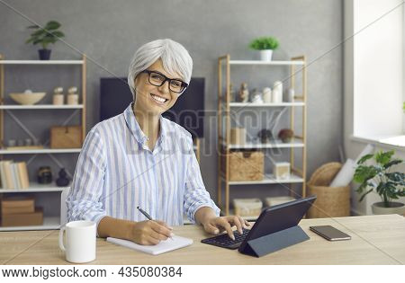 Senior Business Woman Sitting At Desk, Using Tablet Computer And Writing In Notebook