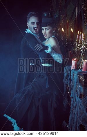 Frightening bloodthirsty male vampire in an elegant suit is going to bite an innocent young waman in his old castle holding her in his arms. Halloween. Vampires of the 19th century. 