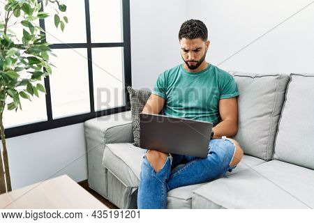 Young handsome man with beard using computer laptop sitting on the sofa at home relaxed with serious expression on face. simple and natural looking at the camera. 