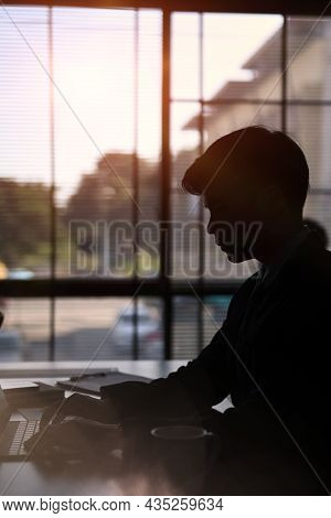 Silhouettes Of Businessman Sitting Near Window And Cocentrate Working With Computer Tablet.
