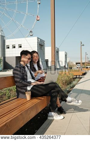 Two young Asian emloyees with laptop preparing presentation while sitting on bench