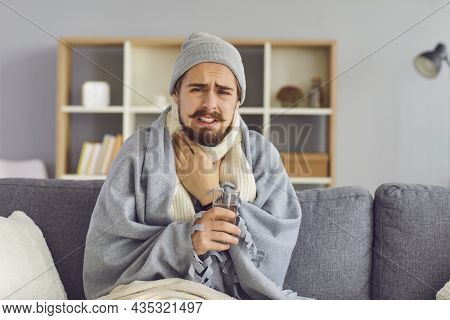 Young Man In Warm Clothes And Blanket Sitting On Sofa With Glass Of Water, Touching Throat And Feeli