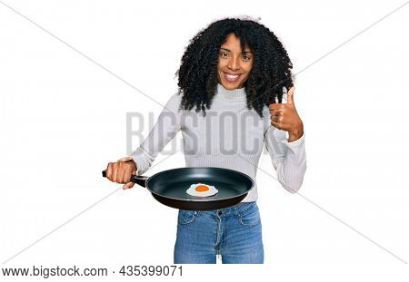Young african american girl holding skillet with fried egg smiling happy and positive, thumb up doing excellent and approval sign 