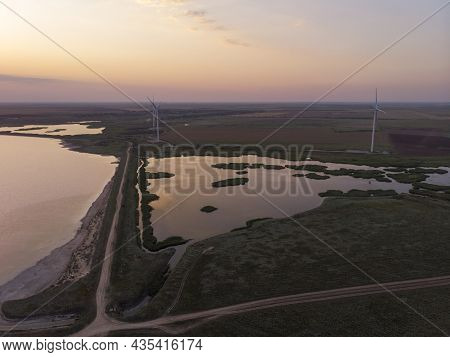 Aerial Drone Shot Of Colorful Pink Lake At Sunset Time With Wind Turbines Farm On Background. Cinema