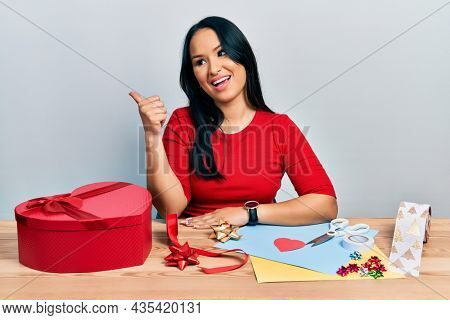 Beautiful hispanic woman with nose piercing doing handcraft creative decoration smiling with happy face looking and pointing to the side with thumb up. 