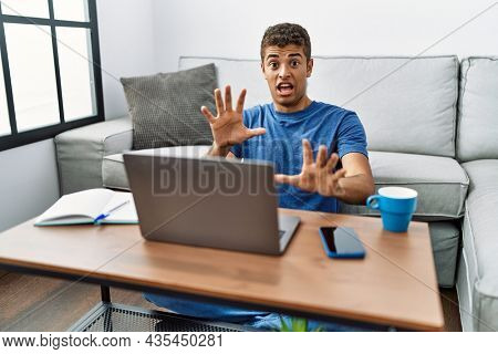 Young handsome hispanic man using laptop sitting on the floor afraid and terrified with fear expression stop gesture with hands, shouting in shock. panic concept. 