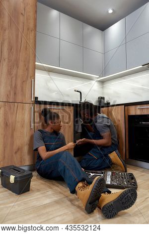 Skilled Male Plumber Checking Pipes Under Kitchen Sink, Female Apprentice Sitting Near, Passing Tool