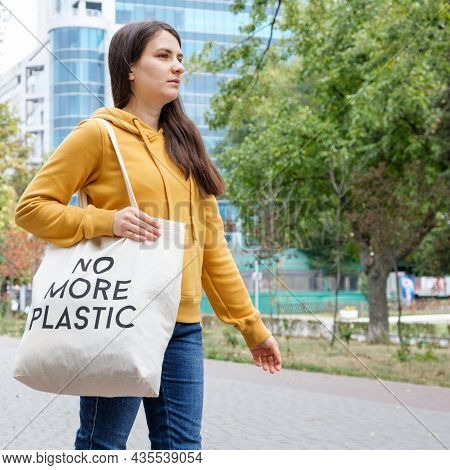 A Brunette Woman In An Orange Hoodie Walks With A Fabric Bag That Says No More Plastic In The City