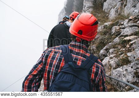 A Young Man In Mountaineering Equipment Walks Uphill On A Suspension Bridge In The Fog. Via Feratta 