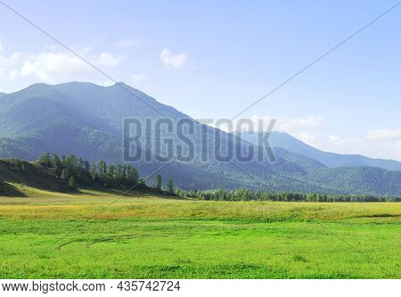 A Green Meadow At The Foot Of The Mountains Under A Blue Cloudy Sky. Siberia, Russia