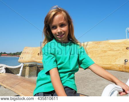 A 7 Years Old White Caucasian Girl In A Green T-shirt Sits On A Wooden Bench, Embankment Of Lake Pal