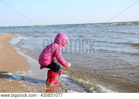 Child Girl Playing By The Sea In Autumn. A Girl In A Red Jacket In A Beautiful Autumn On The Seashor