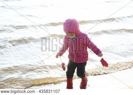 Child Girl Playing By The Sea In Autumn. A Girl In A Red Jacket In A Beautiful Autumn On The Seashor