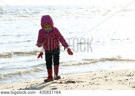 Child Girl Playing By The Sea In Autumn. A Girl In A Red Jacket In A Beautiful Autumn On The Seashor